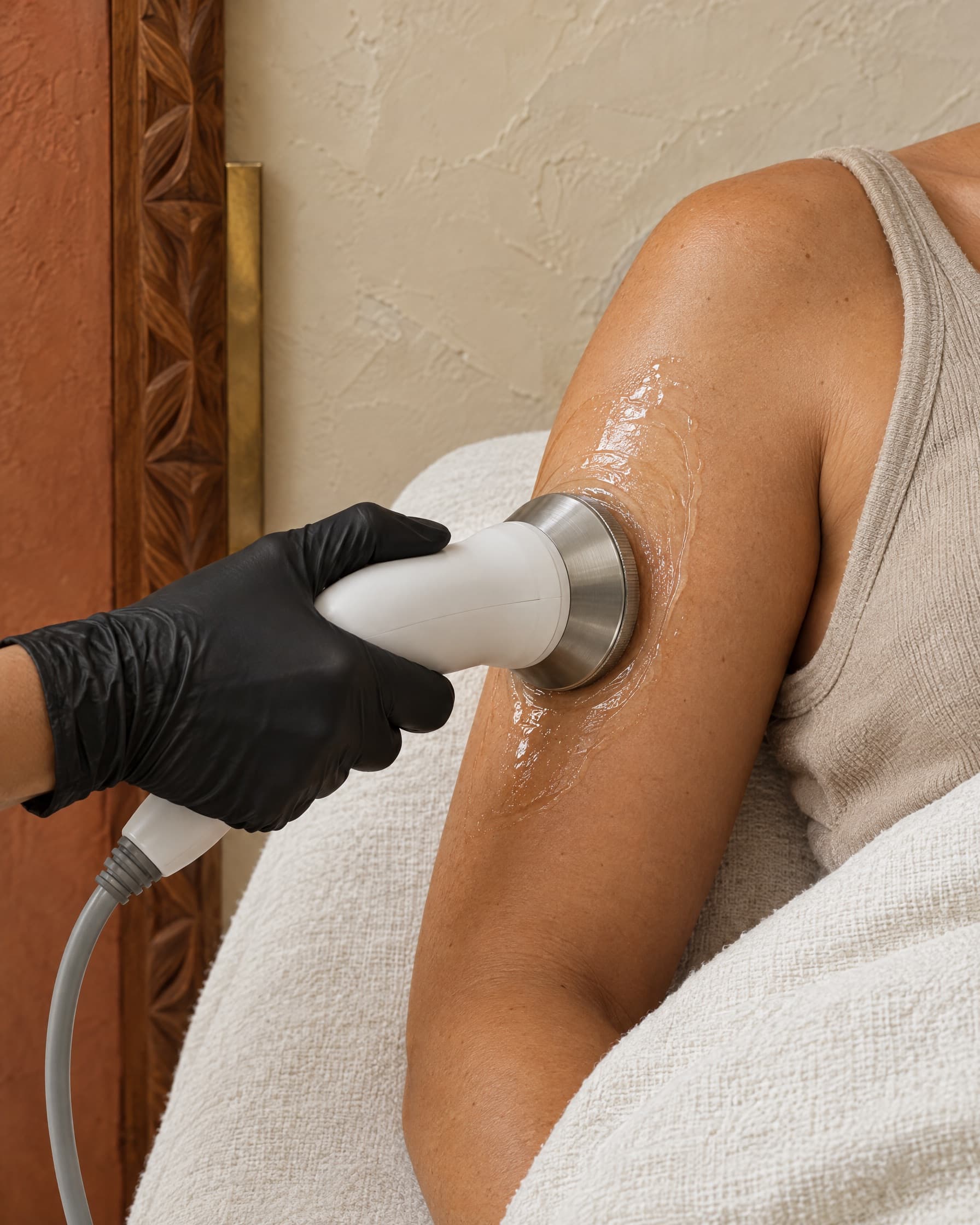 A black-gloved hand moving an ultrasonic cavitation handpiece over gel on the back of a client's upper arm