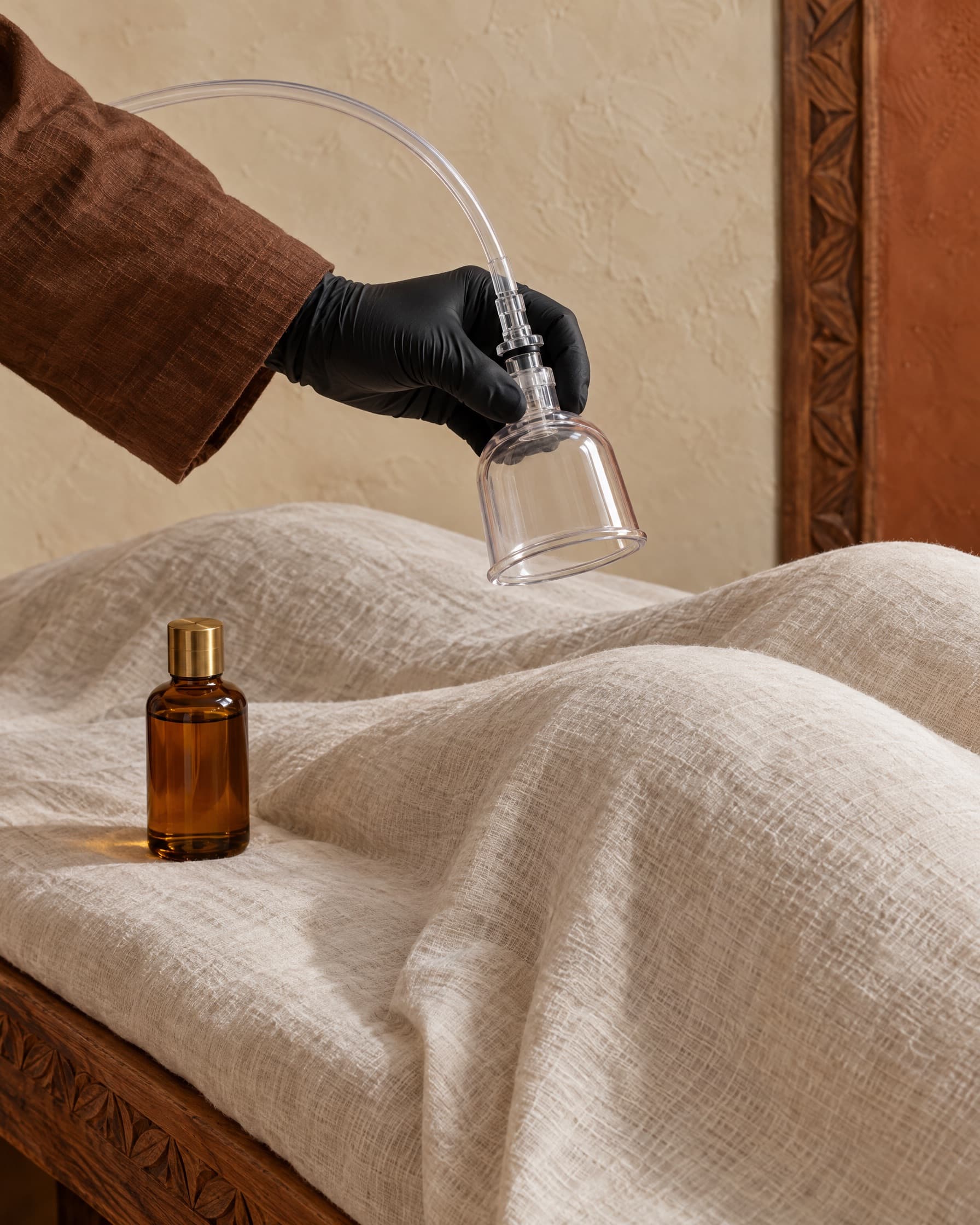 A black-gloved hand holding a clear vacuum therapy cup on tubing above a linen-draped client, a bottle of massage oil beside it
