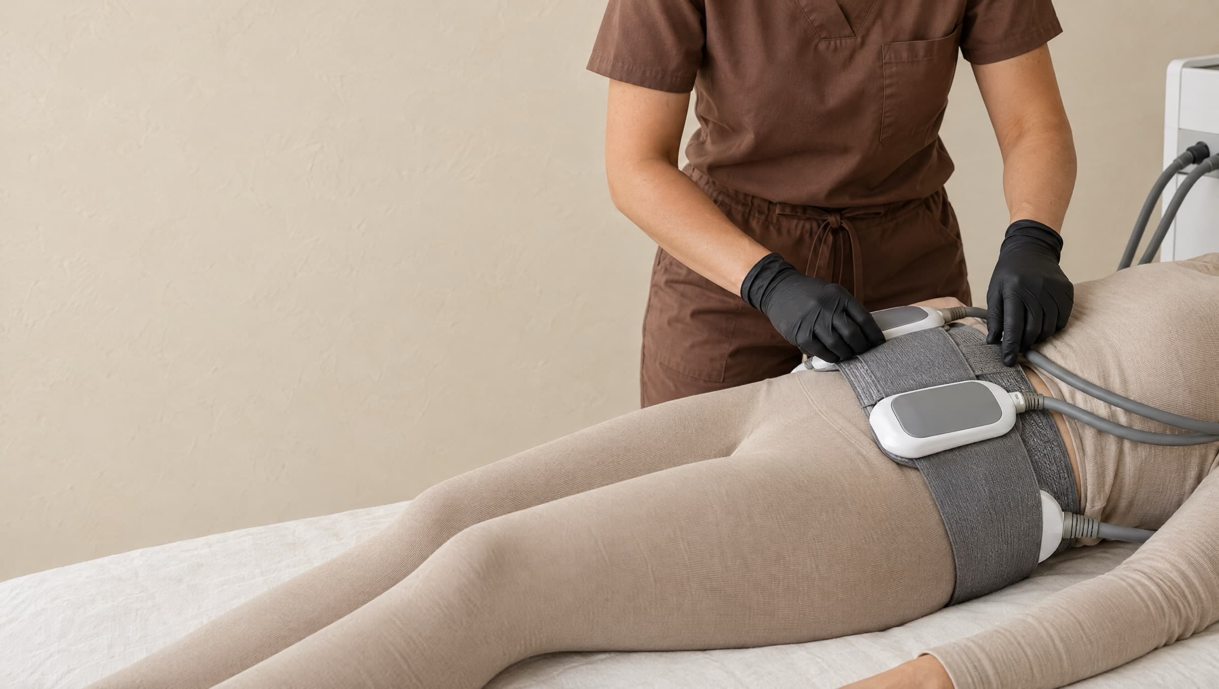 A practitioner's black-gloved hands adjusting the belt that holds two EMS paddles across the midsection of a client lying on a linen-covered treatment bed