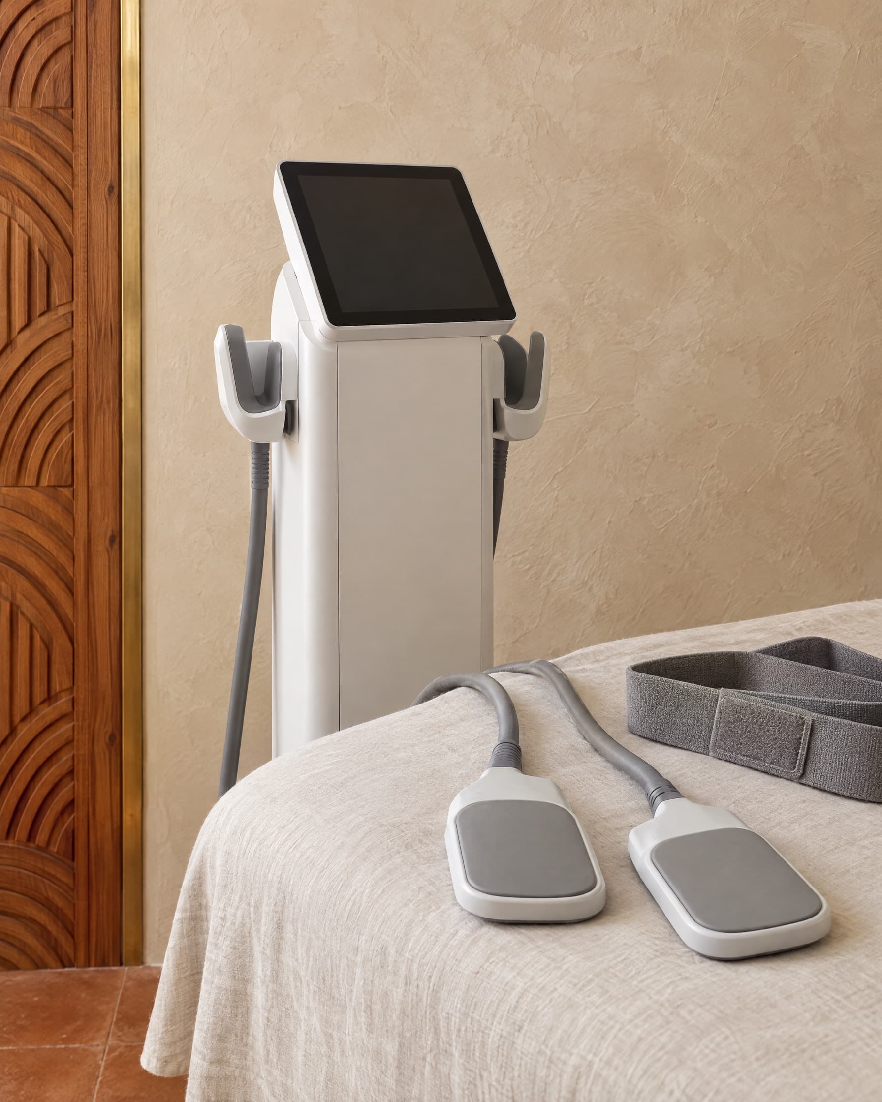 A white EMS tower beside a treatment bed, with two applicator paddles and a grey belt on linen