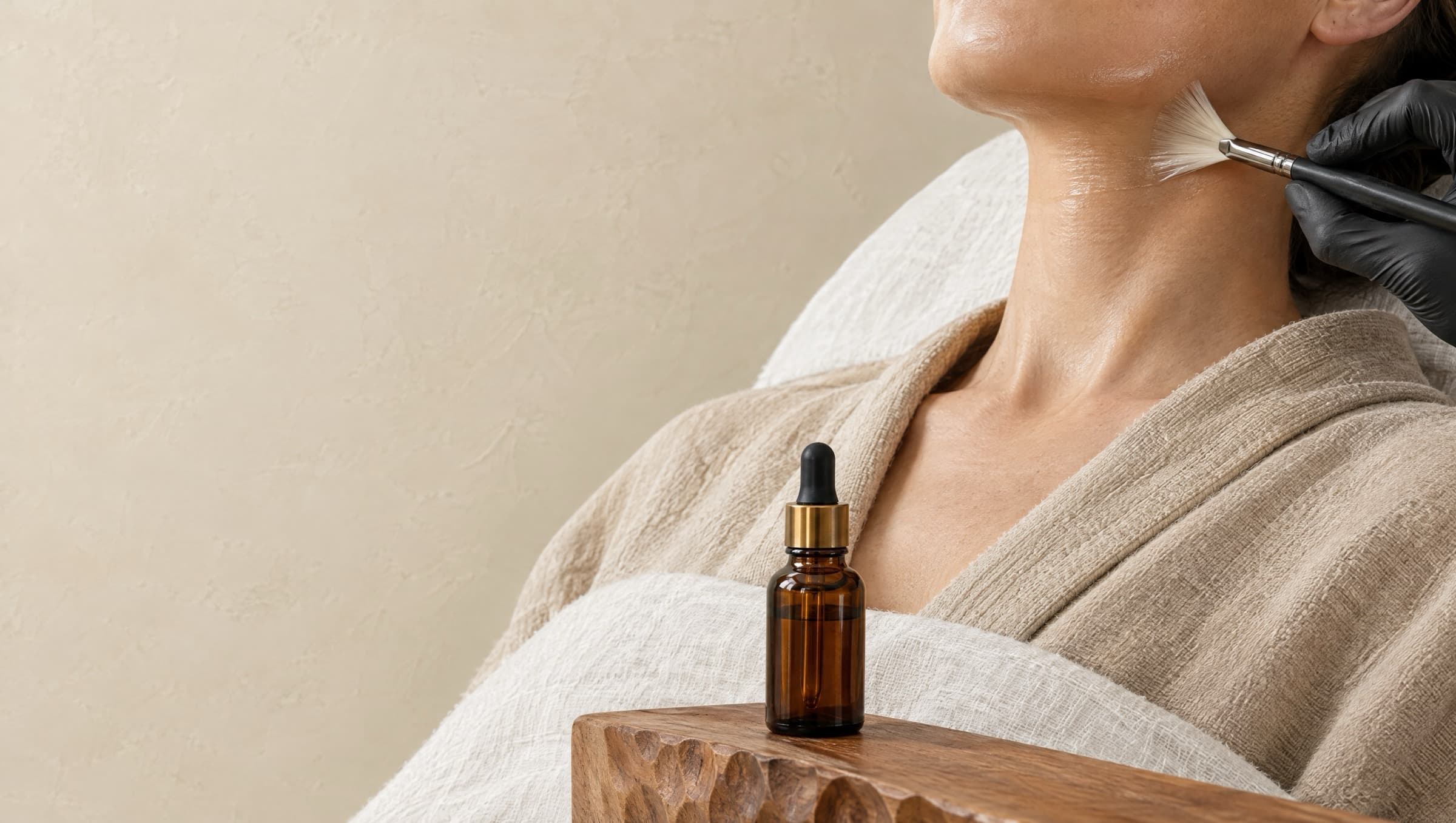 A black-gloved hand sweeping serum along a client's jawline with a fan brush, beside an amber dropper bottle