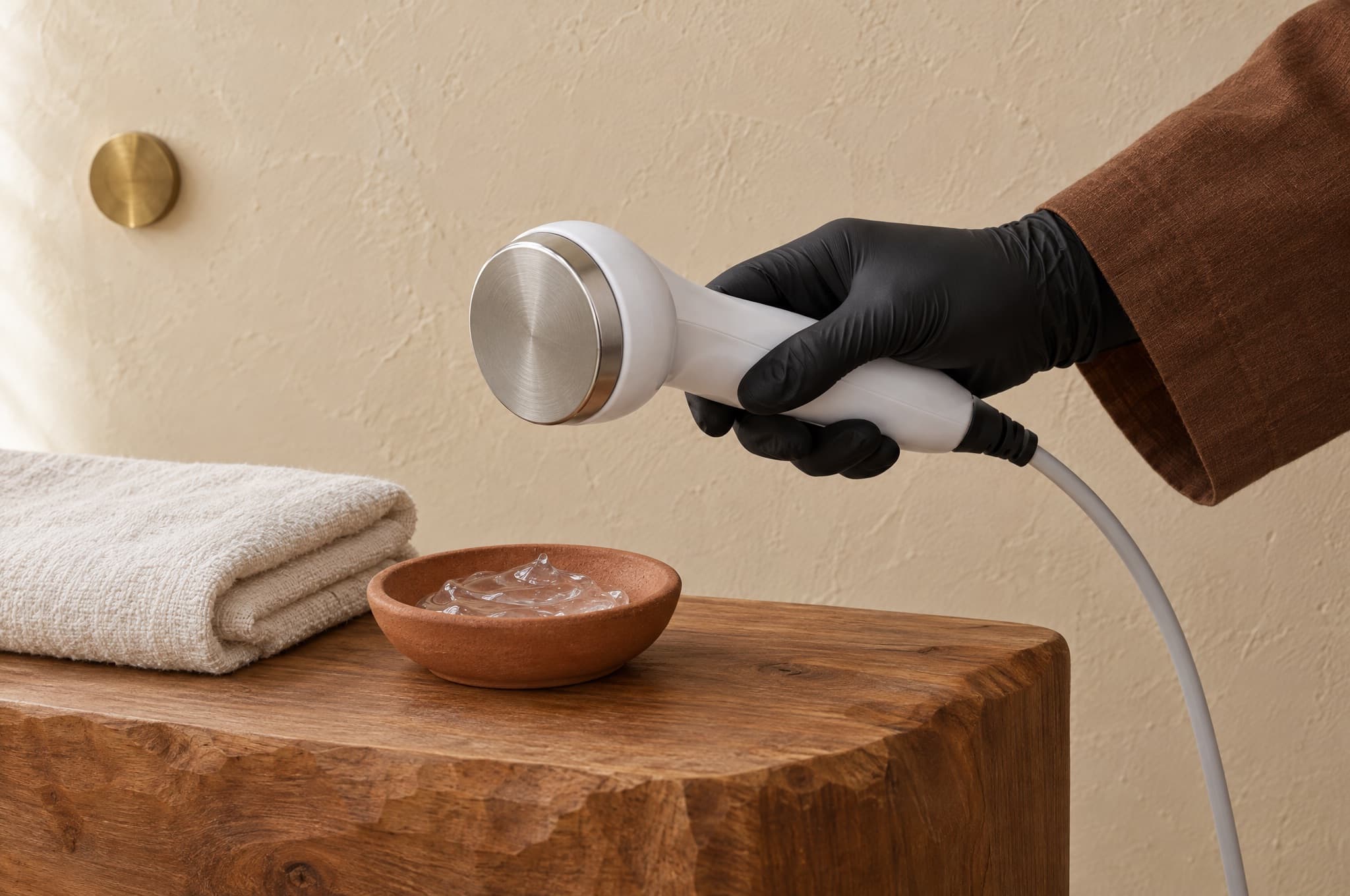 A black-gloved hand holding a cavitation handpiece above a towel and a clay dish of gel