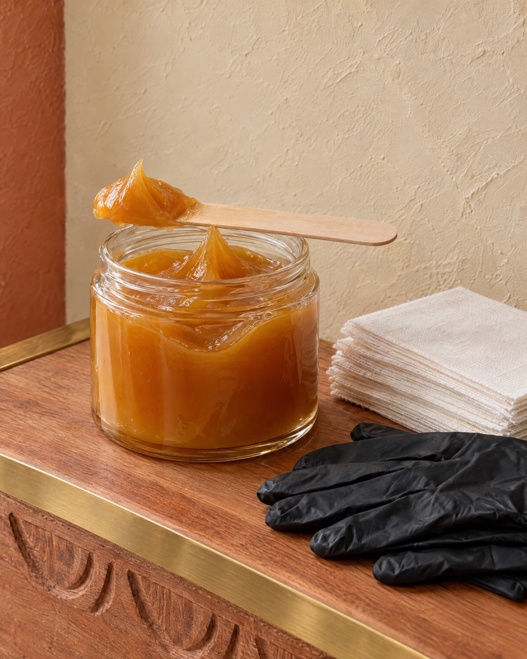 An open jar of amber sugaring paste with a wooden spatula, cotton strips and black gloves on a cedar ledge